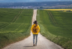 man wearing jeans, a yellow coat and a hat, on a journey on a long path with fields either side.