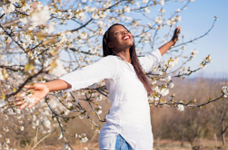 woman standing up, with arms outstretched, big smile standing in front of a blossom tree