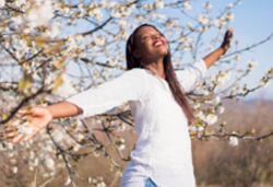 woman standing up, with arms outstretched, big smile standing in front of a blossom tree